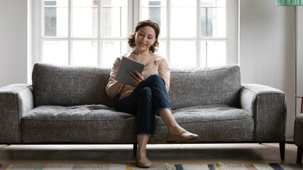 Smiling middle aged woman using tablet, surfing internet, sitting on cozy couch at home, happy mature female enjoying leisure time weekend with gadget, browsing apps, chatting or shopping online
