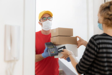 Delivery guy with a medical mask with packages and woman paying with contactless credit card at home during virus pandemic.