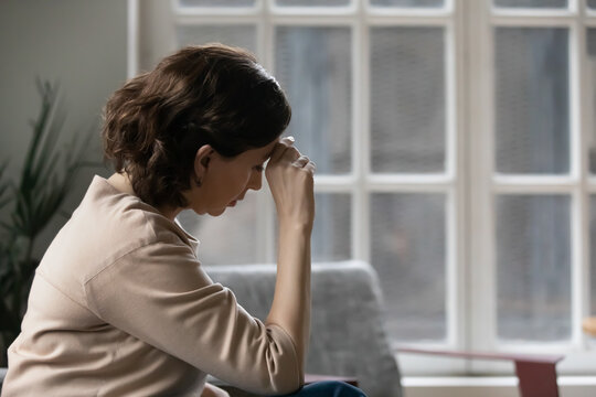 Rear view close up thoughtful mature woman touching forehead, suffering from headache, sitting on couch at home alone, pensive upset middle aged female lost in thoughts, thinking about problems