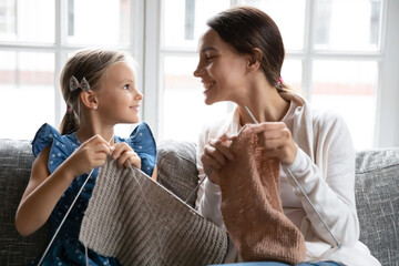 Close up smiling mother and little daughter knitting together, sitting on cozy couch at home, holding needles, happy mum with adorable girl child enjoying leisure time, engaged in creative activity