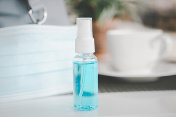 bottle of alcohol spray and blurred surgical mask and white coffee cup, lady bag on white table in open air restaurant , new normal concept after loosen covid-19 lockdown