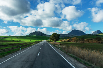 Secondary road that invites the traveler to enjoy the trip between green fields with mountains in the background and a blue sky with cotton clouds.
