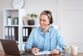 Happy young woman in headset participating in online lesson or web conference, taking notes, studying remotely - obrazy, fototapety, plakaty