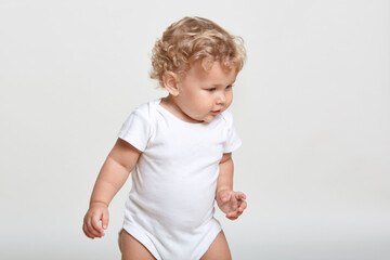 Cute baby boy standing on floor, looking away, sees something interesting, wearing white bodysuit, beautiful tot with blond wavy hair, posing isolated over light background.