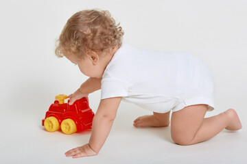 Kid boy toddler playing with toy car, posing on all fours isolated over white background, tot with blond wavy hair playing with red plastic lorry, child wearing bodysuit.