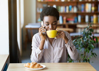Pretty black lady talking on cellphone while drinking coffee at cozy cafe