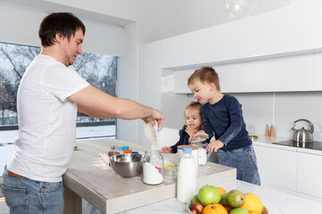 Fototapeta premium A young dad with two young children is preparing pastries in a bright kitchen. Fun pastime. Love, tenderness and care.