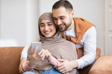 Happy Muslim Couple Using Smartphone Texting Sitting On Couch Indoors