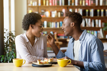 Attractive young black guy feeding his girlfriend with cake, having romantic Valentine's dinner at cozy cafe
