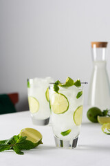 Refreshing cold drinks with lime and mint, glasses with lemonade and ice, jug with drink on white background