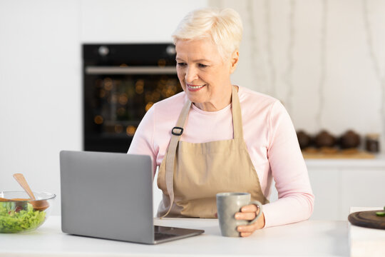 Senior Housewife Using Laptop Browsing Internet Cooking In Kitchen