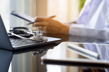 Female doctor hand holding clipboard and work on laptop computer with digital tablet on the table at office in hospital. Online medical, emr, telemedicine, telehealth concept.