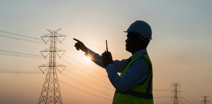 An Asian male engineer using a radio station is working in front of a high voltage pole. In the sunset the sky is beautiful