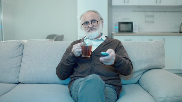 Aged man watching tv at home, drinking tea on sofa, relaxing in free time, hobby