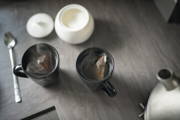 Close up of two black mugs with steaming water and teabags beside a boiling kettle, a spoon and a sugar bowl on a kitchen top in a House in Edinburgh, Scotland, United Kingdom