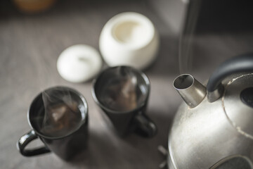 Close up of two black mugs with steaming water and teabags inside beside a boiling kettle, a spoon and a sugar bowl on a kitchen top in a House in Edinburgh, Scotland, United Kingdom