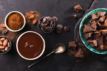 Chunks of dark chocolate on a plate, melted chocolate in a bowl and cocoa beans on a dark textured...