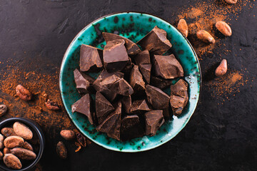 Broken pieces of dark chocolate in a vintage bowl and cocoa beans on a dark textured background, top view. Confectionery chocolate background. Chunks of chocolate.