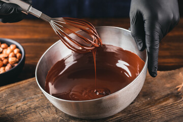 Hand stirring with a whisk melted dark chocolate in a metal bowl on a wooden table. Chocolate...