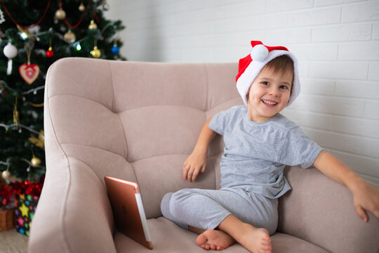 Joyful child wears santa hatlooking at the camera at christmas while sitting in a chair in the living room at home, holding a tablet