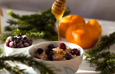 muesli in a white plate on the table, in which honey flows from a spoon on top. Frozen berries lie...