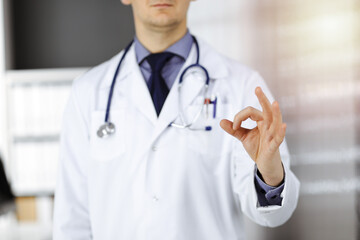 Unknown male doctor standing straight with OK sign in sunny clinic, closeup. Perfect medical service in hospital. Medicine and healthcare concept