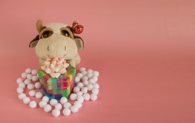 The symbol of 2021 year, a soft toy cow with a gift box and decorative snowballs on a pink background. Christmas and New Year postcard. Copy space