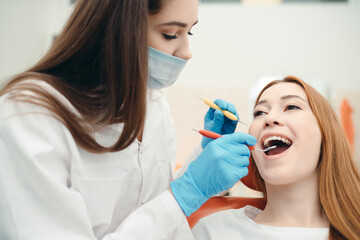A masked brunette examines a client's teeth using special medical devices