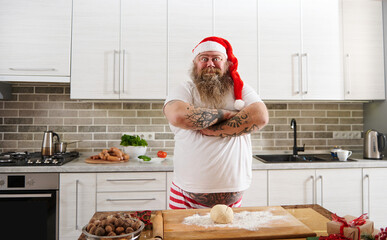 An overweigh man in Santa Claus hat standing in front of the table in the kitchen and looking at camera with his tattooed arms crossed.