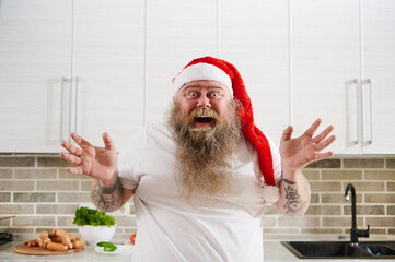 A smiling funny overweigh man in Santa Claus hat with tattooed arms wearing a white T-Shirt looking at camera thereby showwing his emotions: surprise, excitation, happiness