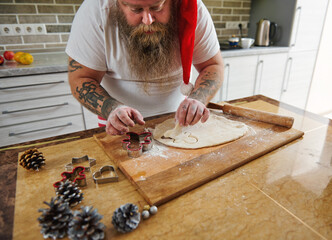 A bearded man wearing Santa Claus hat with tattooed arms cutting a shape out of dough and passionate about cooking gingerbread. Pines cones and cookie molds on the table in the kitchen.