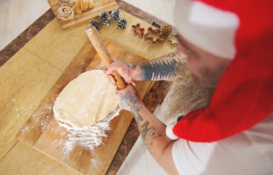 A Top View Of A Man In A Red Santa Hat And Tattooed Arms Holding A Wooden Rolling Pin Like A Baseball Bat. A Flat Dough And Some Christmas Decorations On A Table In The Kitchen