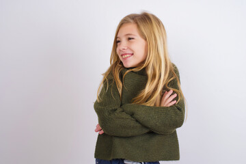 Dreamy rest relaxed Caucasian kid girl wearing green knitted sweater against white wall crossing arms, looks good copyspace