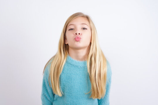 Cute Caucasian kid girl wearing blue knitted sweater against white wall, keeps lips as going to kiss someone, has glad expression, grimace face. Standing indoors. Beauty concept.