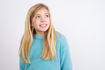 Fototapeta premium Oops! Portrait of Cute Caucasian kid girl wearing blue knitted sweater against white wall clenches teeth and looks confusedly aside, realizes her bad mistake,