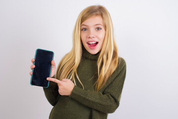 Attractive cheerful Caucasian kid girl wearing green knitted sweater against white wall holding in hands cell showing black screen