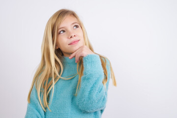 Dreamy Cute Caucasian kid girl wearing blue knitted sweater against white wall with pleasant expression, looks sideways, keeps hand under chin, thinks about something pleasant.