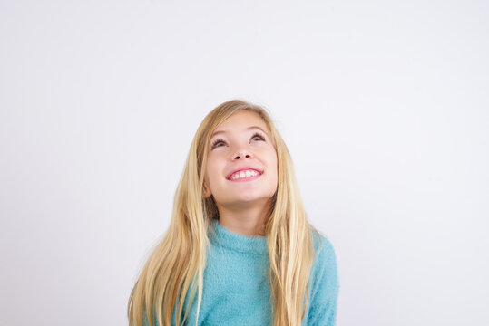 Portrait of mysterious Cute Caucasian kid girl wearing blue knitted sweater against white wall looking up with enigmatic smile. Advertisement concept.
