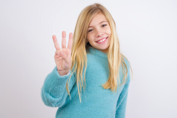 Cute Caucasian kid girl wearing blue knitted sweater against white wall showing and pointing up with fingers number three while smiling confident and happy.