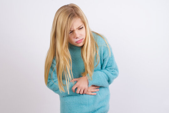 Cute Caucasian kid girl wearing blue knitted sweater against white wall suffering from strong stomachache.