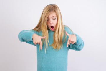 Amazed Cute Caucasian kid girl wearing blue knitted sweater against white wall points down with...