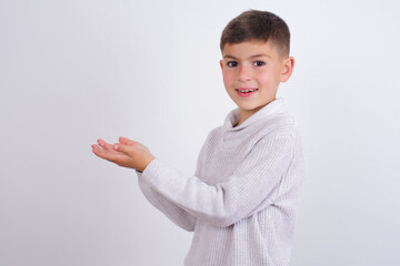 Cute Caucasian kid boy wearing knitted sweater against white wall pointing aside with hands open...