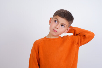 Cute Caucasian kid boy wearing knitted sweater against white wall confuse and wonder about question. Uncertain with doubt, thinking with hand on head. Pensive concept.
