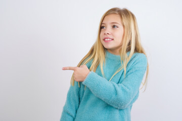 Happy cheerful smiling Cute Caucasian kid girl wearing blue knitted sweater against white wall looking and pointing aside with hand. Copy space and advertisement concept.