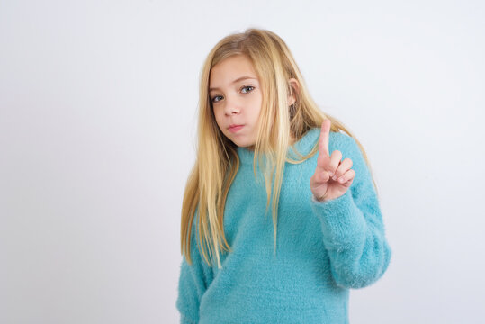 No sign gesture. Closeup portrait unhappy Cute Caucasian kid girl wearing blue knitted sweater against white wall raising fore finger up saying no. Negative emotions facial expressions, feelings.