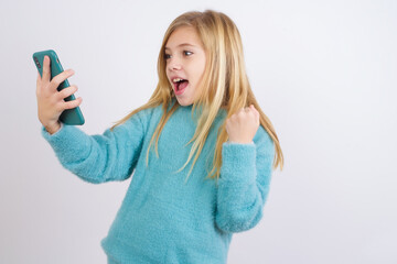 Happy cheerful Cute Caucasian kid girl wearing blue knitted sweater against white wall receiving good news via e-mail and celebrating success while standing and looking at mobile phone.