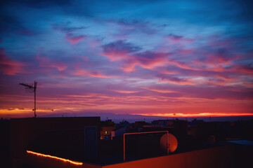 Sun appears from behind the clouds, above the city, roof top view. Summer sunrise or sunset. Dark blue and red sky color tones. Old town of Spain.