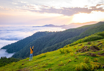 Female person out of joy looks to camera with spreaded hands and scenic landscape of Gomismta mountain above the clouds with sunrise in backgorun