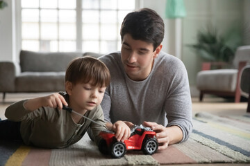 Close up father and little son playing, fixing toy car, lying on warm floor with underfloor heating...