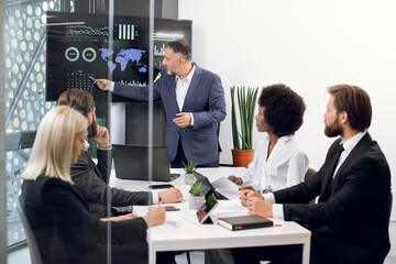 Team of diverse attentive mixed race business people listening their middle aged male bearded chief, showing dynamics of key ecological global problems, or world market, using digital wall screen